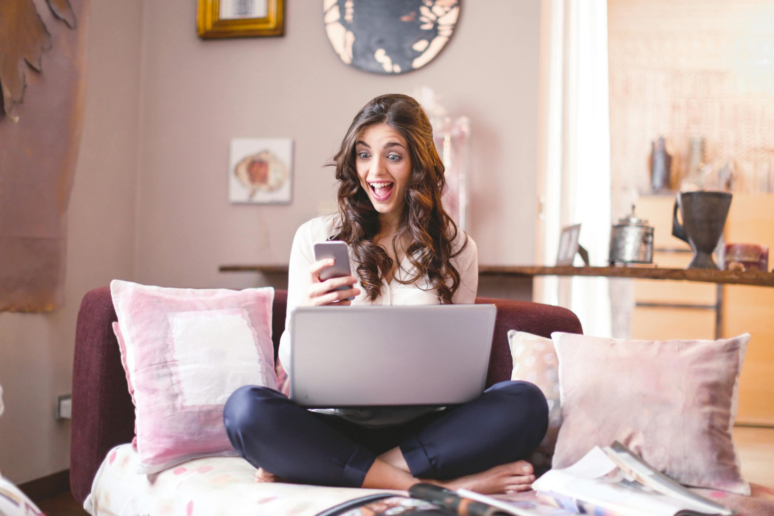 Smiling woman at home, using smartphone and laptop, expressing surprise and happiness.