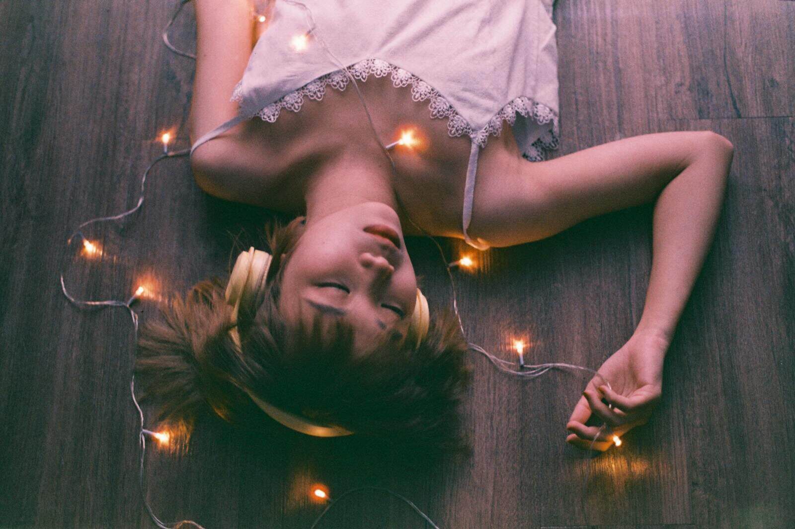 Young woman lies on wooden floor with fairy lights and headphones, eyes closed.