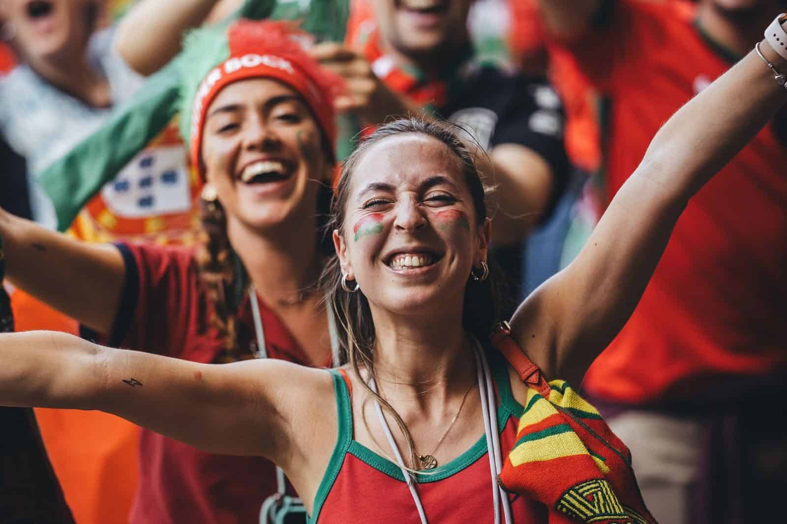 Excited women fans celebrate with flags and smiles during a sports event outdoors.