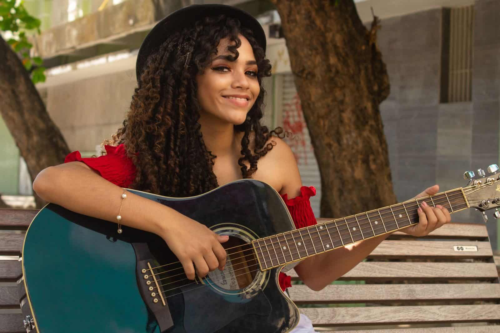 Smiling young woman in a red top playing guitar on a bench outdoors in Brazil.
