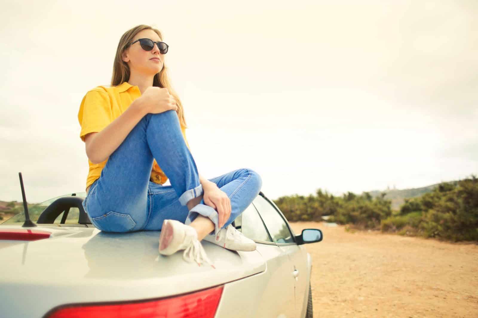 Young woman wearing sunglasses sits on car roof, enjoying a sunny day in Malta.