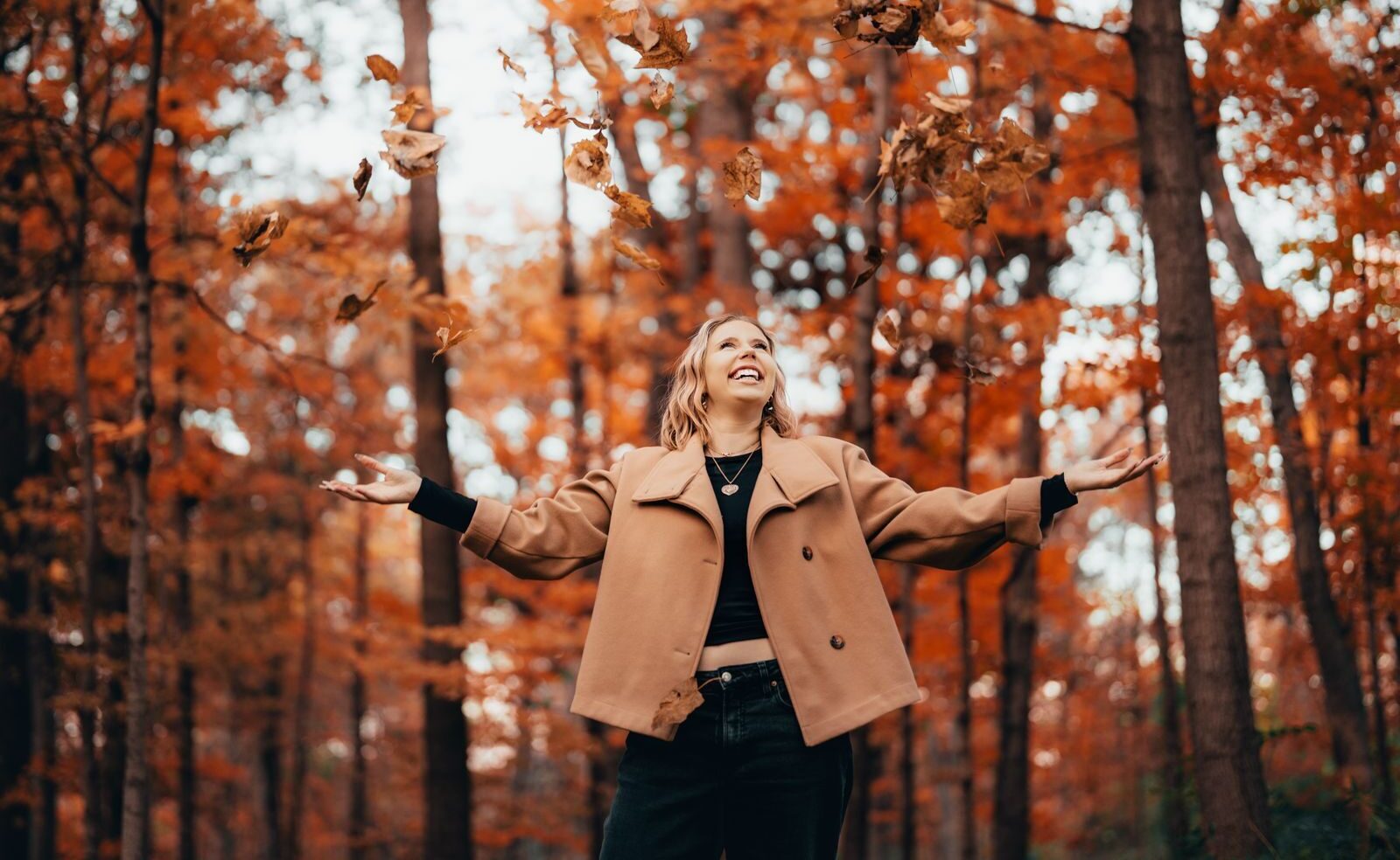 A woman joyfully throws leaves in a vibrant autumn forest, surrounded by orange foliage.